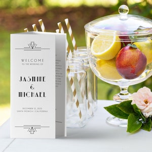 May include: A white wedding welcome sign with black text that reads "Welcome to the wedding of Jasmine & Michael December 12, 2023 Santa Monica | California". The sign is decorated with a geometric design.