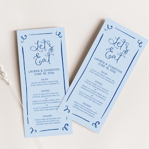 May include: Two light blue wedding menu cards with a navy blue border and calligraphy that says "Let's Eat". The cards list the menu for a wedding reception with the names Lauren & Jonathon and the date June 18, 2026. The menu includes salad, entree, and dessert options.