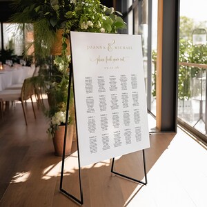 May include: A white wedding seating chart with black text on an easel stand. The chart lists tables and guest names for Joanna & Michael's wedding on 09.12.16.  The text reads "Please find your seat."