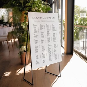 May include: A white wedding seating chart with a black stand. The chart lists guest names alphabetically by table. The chart is titled "Samara and Carlos" with the date "September 27, 2026" and location "Carmel, California".