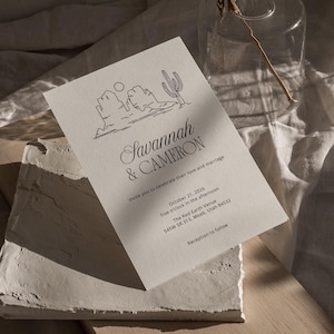 Può includere: Un invito di nozze minimalista con un disegno a linea nera di un paesaggio desertico con un cactus e montagne. Il testo recita "Savannah & Cameron vi invitano a celebrare il loro amore e matrimonio il 21 ottobre 2026 alle cinque del pomeriggio presso The Red Earth Venue 545W SR 313. Moab, Utah 84532 Ricevimento a seguire"