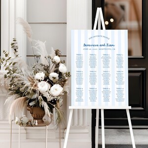 Könnte beinhalten: Eine weiße Staffelei zeigt einen Sitzplan mit hellblauem Streifenmuster. Die Tabelle listet Namen unter Tischnummern auf, mit dem Text "OUR FAVORITE PEOPLE" und "Genevieve and Liam" oben. Datum und Ort sind ebenfalls angegeben.
