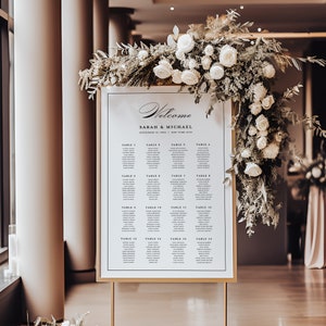 Könnte beinhalten: Ein weißer Hochzeitssitzplan mit goldenem Rahmen und goldenem Ständer. Der Plan listet die Namen der Gäste auf, die den einzelnen Tischen zugeordnet sind. Der Plan ist mit weißen Blumen und Grünpflanzen dekoriert.