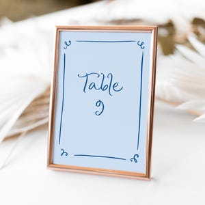 May include: A rose gold frame with a light blue background and a navy blue inscription that reads "Table 9".