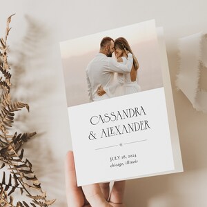 May include: A folded wedding invitation featuring a photograph of a couple embracing. The invitation is white with the names "Cassandra & Alexander" in elegant black script. The date "July 18, 2024" and location "Chicago, Illinois" are also printed.
