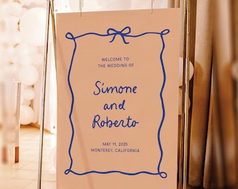 May include: A peach-colored wedding sign with a blue border and a blue bow at the top. The sign reads "Welcome to the wedding of Simone and Roberto, May 11, 2025, Monterey, California."