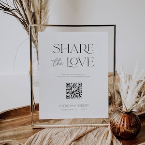 May include: A white sign with a gold frame, with the text "SHARE the LOVE" in black. Below the text is a black and white QR code with the text "Please share your photos with us. Simply scan with your smartphone and upload." Below the QR code is the text "ASTRID and ROBERT FEBRUARY 21, 2022".