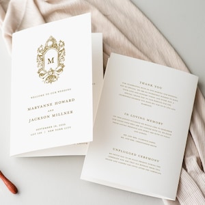 May include: Ivory wedding program with gold foil accents. The front features a decorative frame with the letter "M" and the words "Welcome to our Wedding." Inside, text includes "Thank You" and "In Loving Memory."