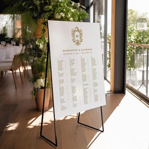 Könnte beinhalten: Ein weißer Hochzeitssitzplan mit goldenen Buchstaben auf einem Staffelei. Der Sitzplan listet die Namen der Gäste alphabetisch nach Tisch auf. Oben auf dem Sitzplan befindet sich ein goldenes Wappen mit dem Buchstaben "M" und den Namen "Maryanne & Jackson" sowie dem Datum "18. September 2023" und dem Ort "New York City".