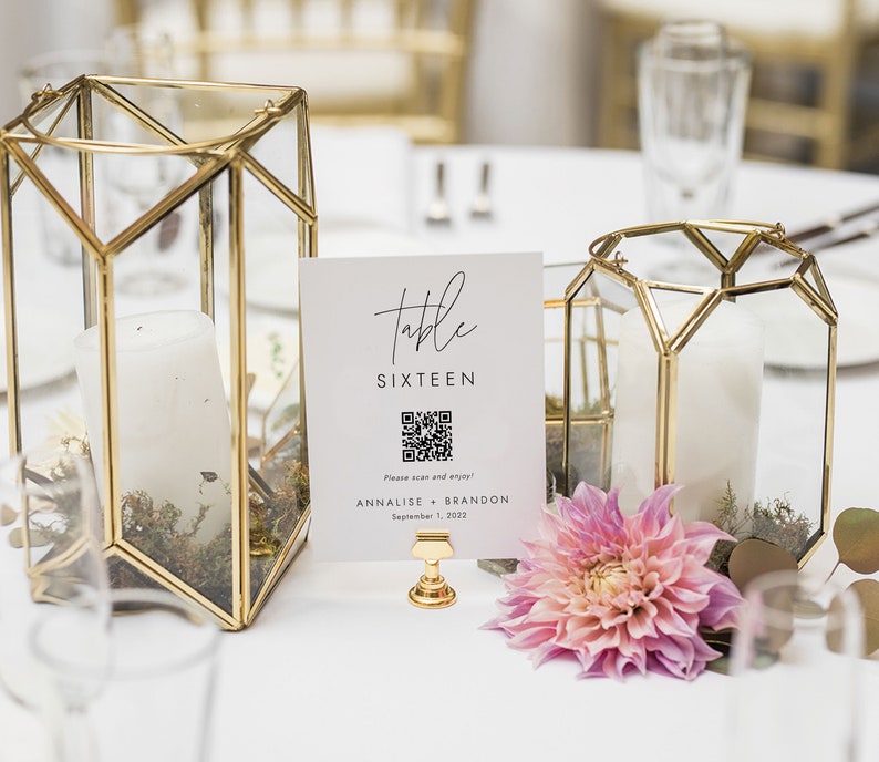 Op de afbeelding: Een witte tafelnummerkaart met de tekst "Table Sixteen" en een QR-code. De kaart staat op een gouden standaard en heeft een roze bloem ervoor. De kaart bevat ook de namen "Annalise + Brandon" en de datum "1 september 2022".