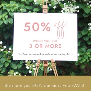 May include: A white sign with pink text announcing a 50% off sale when you buy 3 or more items. The sign is on a wooden easel in front of a green bush with white flowers.