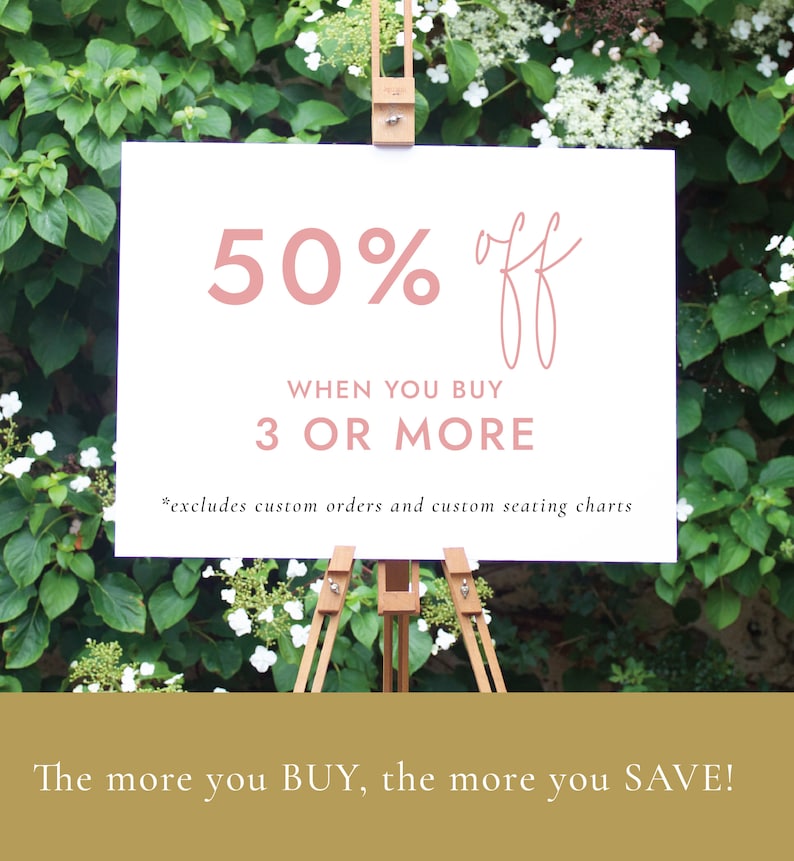May include: A white sign with pink text announcing a 50% off sale when you buy 3 or more items. The sign is on a wooden easel in front of a green bush with white flowers.