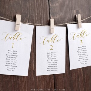 Könnte beinhalten: Drei weiße Karten mit goldener Kalligraphie, auf denen "Tisch 1", "Tisch 2" und "Tisch 3" steht. Jede Karte listet eine Gruppe von Namen für einen Hochzeitsempfang-Sitzplan auf.