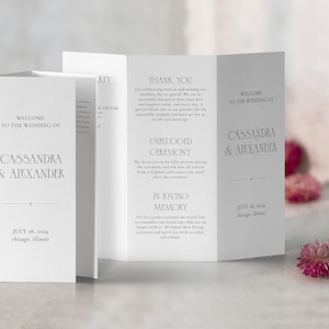 May include: A white wedding program with a simple design. The front cover reads "Welcome to the Wedding of Cassandra & Alexander" with the date "July 18, 2024" and location "Chicago, Illinois". The inside pages include a "Thank You" message, an "Unplugged Ceremony" request, and an "In Loving Memory" section.