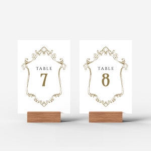 K&ouml;nnte beinhalten: Zwei wei&szlig;e Tischnummernkarten mit goldenen, verzierten Rahmen und schwarzem Text. Die Karten stehen auf Holzst&auml;ndern. Der Text auf den Karten lautet "Tisch 7" und "Tisch 8".