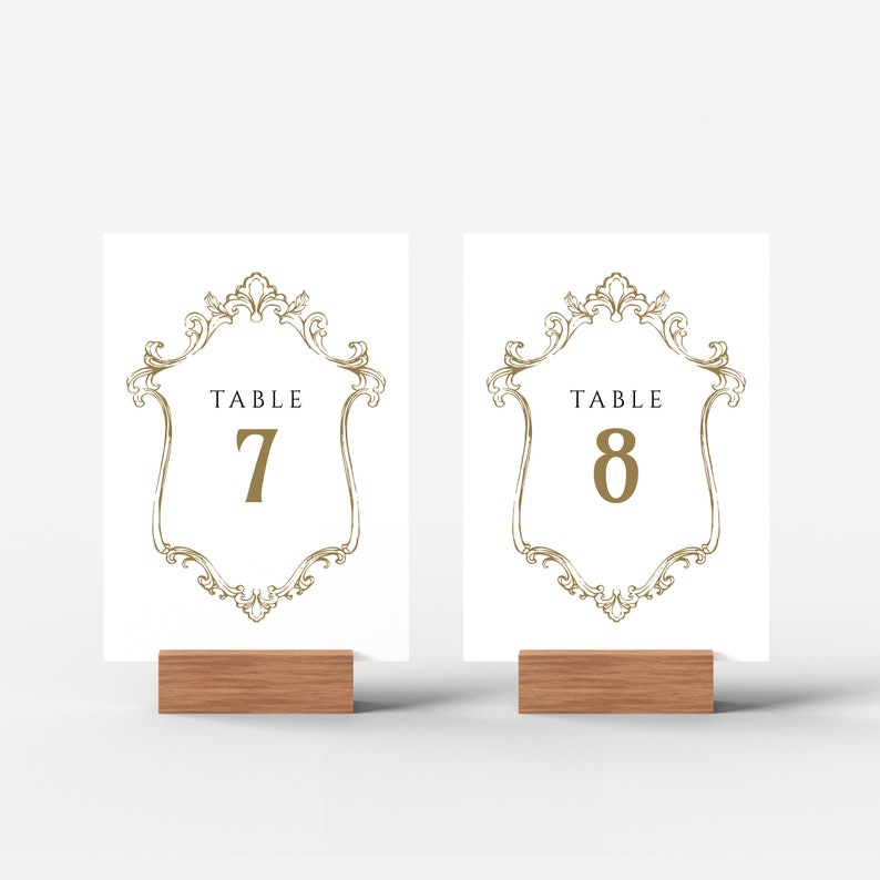 K&ouml;nnte beinhalten: Zwei wei&szlig;e Tischnummernkarten mit goldenen, verzierten Rahmen und schwarzem Text. Die Karten stehen auf Holzst&auml;ndern. Der Text auf den Karten lautet "Tisch 7" und "Tisch 8".