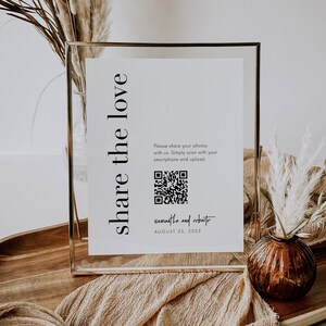 May include: A gold-framed sign with a white background and black text that reads "share the love". A QR code is displayed below the text, along with the names "Samantha and Roberto" and the date "August 23, 2023".
