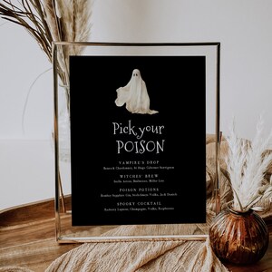 Könnte beinhalten: Ein schwarzes Schild mit dem Text "Pick your POISON" und einer Geisterillustration. Das Schild listet Cocktailnamen und Zutaten auf, darunter "Vampire's Drop" und "Spooky Cocktail". Der Rahmen ist goldfarben.