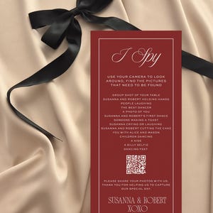 May include: A burgundy "I Spy" wedding game card with white text, featuring a list of photo prompts. A black satin ribbon and bow are draped across a cream-colored satin background.