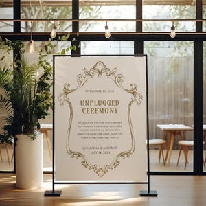May include: A white sign with a black frame and gold ornate design. The sign reads "Welcome to our Unplugged Ceremony" and includes the date "July 18, 2025".