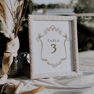 K&ouml;nnte beinhalten: Wei&szlig;e Tischnummerkarte mit einem goldenen verzierten Rahmen und dem Text "TABLE 3" in goldenen Buchstaben.