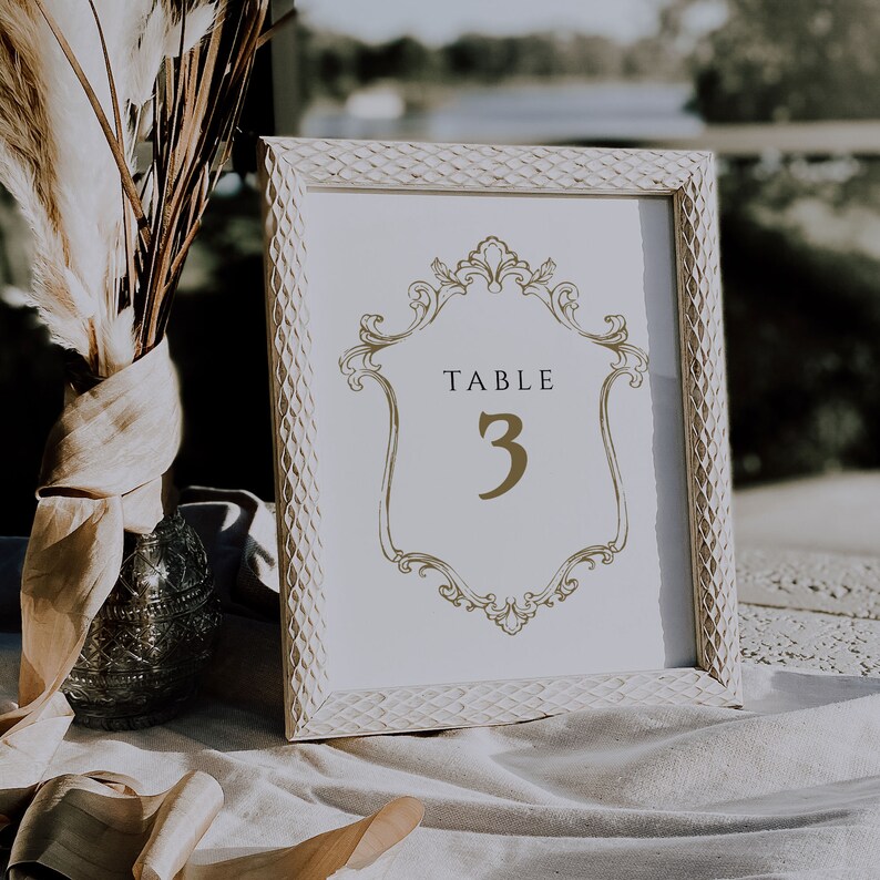 K&ouml;nnte beinhalten: Wei&szlig;e Tischnummerkarte mit einem goldenen verzierten Rahmen und dem Text "TABLE 3" in goldenen Buchstaben.