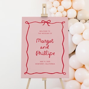 May include: Pink wedding welcome sign with a red string border and bow. The sign reads "Welcome to the wedding of Margot and Phillipe, May 11, 2025, Monterey, California."
