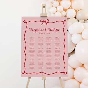 Könnte beinhalten: Ein rosa Hochzeits-Sitzplan auf einer Staffelei mit roter Schleife und Rahmendesign. Die Namen "Margot und Phillipe" und das Datum "11. Mai 2025" sind in einer dekorativen Schriftart dargestellt. Die Gästenamen sind unter den Tischnummern aufgeführt.
