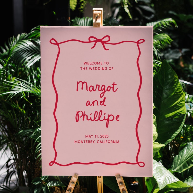 May include: Pink wedding sign with a red ribbon and a handwritten message that says "Welcome to the wedding of Margot and Phillipe, May 11, 2025, Monterey, California."