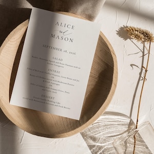 Op de afbeelding: Een huwelijksmenukaart met een minimalistisch ontwerp. De kaart is wit met zwarte tekst en bevat de namen "Alice en Mason" en de datum "28 september 2026". Het menu bevat een salade, een hoofdgerecht en een dessert.