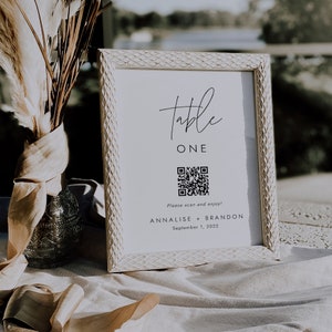 Op de afbeelding: Een wit tafelnummerbord met een zwarte lijst. Het bord zegt "Table One" en heeft een QR-code met de tekst "Please scan and enjoy!"  Het bord zegt ook "Annalise + Brandon September 1, 2022".