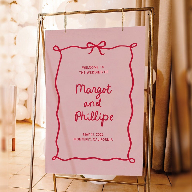 May include: Pink wedding welcome sign with a red ribbon and a red border. The sign reads "Welcome to the wedding of Margot and Phillipe, May 11, 2025, Monterey, California."