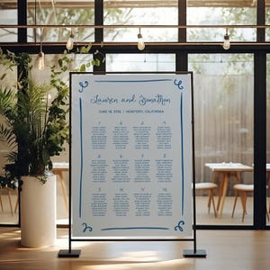 Könnte beinhalten: Ein weißer Hochzeitssitzplan mit blauer Kalligrafie. Der Plan ist mit "Lauren und Jonathan" betitelt und listet das Datum und den Ort der Hochzeit auf. Der Plan ist in 12 Tische unterteilt, die jeweils mit einer Nummer und einer Namensliste versehen sind.