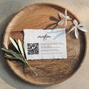 May include: A reception invitation card with a QR code, printed on textured paper. The card is on a wooden plate with white flowers. The text on the card reads: "reception Please join us at Half Past Six in the Evening for Cocktails, Dinner and Dancing The Cliffon Manor 7245 The Esplanade, Santa Ana, CA For further information please SCAN HERE or access our website ELIZABETHANDMAX.COM"