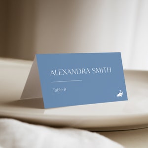 以下が含まれることがあります： 白い文字で「ALEXANDRA SMITH」と「Table 8」と書かれた青い席札。右下に白い魚のシルエットがあります。