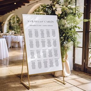 Könnte beinhalten: Eine weiße Hochzeits-Sitzordnung mit goldenem Metallrahmen. Die Tafel zeigt die Namen der Gäste, geordnet nach Tischnummer. Der Text lautet "SAMARA and CARLOS" und "September 27, 2026 | Carmel, California".