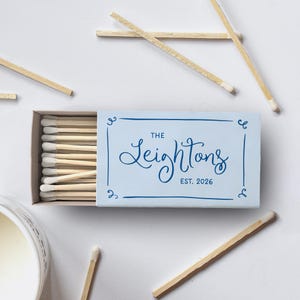 Puede incluir: Una caja de cerillas azul claro con el nombre "The Leightons" y el año "EST. 2026" impresos en azul oscuro. La caja está abierta, revelando una fila de cerillas con puntas blancas. Varias cerillas sueltas están esparcidas alrededor de la caja.