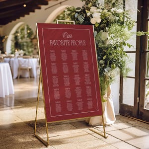 Könnte beinhalten: Ein weinroter Sitzplan mit dem Text "Our Favorite People" in eleganter Schrift. Die Tabelle listet Tischnummern in einem Rasterformat auf, das auf einem goldenen Metallständer angezeigt wird. Der Hintergrund zeigt Blumengestecke und eine verschwommene Inneneinrichtung.