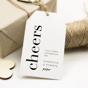 May include: White gift tag with black text that reads "cheers" in a vertical orientation. The text "to love, laughter, and happily ever after" is below the word "cheers". The names "Samantha & Roberto" and "xoxo" are below the text.