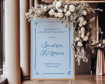 May include: A blue and white sign with gold trim announcing a graduation party for Lauren Kirkman, Class of 2025. The sign is decorated with white flowers and greenery.