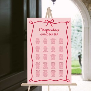 Op de afbeelding: Roze zitplaatskaart met een rode strik en een rode touwrand. De tekst luidt "Margarita's Quinceañera" en geeft een lijst met tafels met namen voor elke tafel.