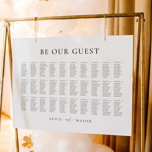 Könnte beinhalten: Ein weißer Sitzplan mit dem Text "Be Our Guest" oben. Der Plan listet die Namen der Gäste nach Tischnummer auf, von Tisch 1 bis Tisch 20. Der Plan wird auf einem goldenen Metallständer angezeigt. Am unteren Rand des Plans steht "Alice & Mason".