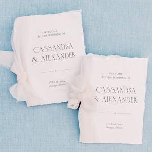 Puede incluir: Dos programas de boda blancos con bordes rasgados, atados con cintas blancas, sobre un fondo azul claro. Los programas dicen "WELCOME TO THE WEDDING OF CASSANDRA & ALEXANDER" con la fecha 18 de julio de 2024 y la ubicación Chicago, Illinois.