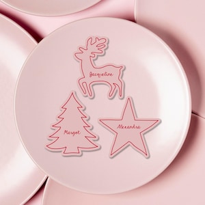 Puede incluir: Un plato rosa con tres tarjetas decorativas. Las tarjetas tienen forma de reno, árbol de Navidad y estrella. La tarjeta de reno tiene el nombre "Jacqueline", el árbol "Margot" y la estrella "Alexandra".