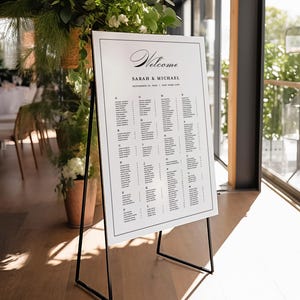 Könnte beinhalten: Ein weißer Hochzeitssitzplan mit schwarzem Rahmen auf einem schwarzen Staffelei-Ständer. Der Plan trägt oben die Worte "Willkommen Sarah & Michael" und listet die Namen der Gäste alphabetisch nach Tischen auf.