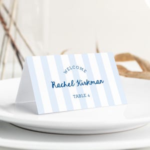 May include: A folded place card with a blue and white striped pattern. The card reads "WELCOME" above the name "Rachel Kirkman" in a script font, with "TABLE 4" below. The card is on a white plate.