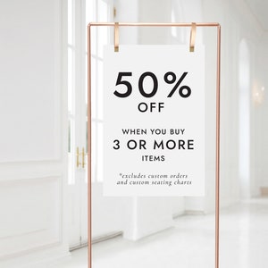 Op de afbeelding: Een wit bord met een koperen frame en clips, dat een 50% korting aankondigt bij aankoop van 3 of meer artikelen. Het bord luidt "50% KORTING BIJ AANKOOP VAN 3 OF MEER ARTIKELEN *exclusief aangepaste bestellingen en aangepaste zitplaatsen".
