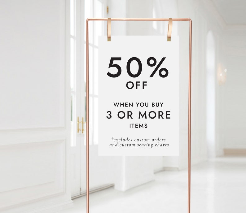 Op de afbeelding: Een wit bord met een koperen frame en clips, dat een 50% korting aankondigt bij aankoop van 3 of meer artikelen. Het bord luidt "50% KORTING BIJ AANKOOP VAN 3 OF MEER ARTIKELEN *exclusief aangepaste bestellingen en aangepaste zitplaatsen".