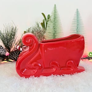 May include: A glossy red ceramic Santa's sleigh planter. The decorative sleigh has a curved front and back, with a rectangular opening for plants. The sleigh is on a snowy surface, with Christmas decorations in the background.