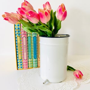May include: A white enamelware pitcher with a black rim and spout, holding pink and green tulips. Behind the pitcher are colorful cookbooks with titles like "Desserts" and "Meats". A white doily is beneath the pitcher.